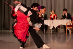 A male dancer in black trousers, white long sleeve top, black vest, and white shoes holds a female dancer in a fish lift. The female dancer curves her left arm in front of her and her right arm extended to the side. She wears a red flamenco-like skirt and black long-sleeve top with a red flower on the side of her bun.