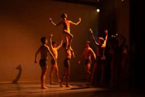 A soft, warm orange light illuminates a dark stage. A group of male dancers in beige long-sleeve shirts and black shorts surround a female dancer in a warm-toned light orange leotard. She is held aloft by one of the men in a torch lift. Her left leg is straight down with a pointed foot, and her right leg is in a parallel coupe position near her ankle.
