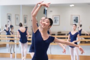 A close up shot of a 2nd year student in a dark blue cap sleeve leotard with her right arm in fifth and her left arm to the side. She smiles to the teacher off camera.