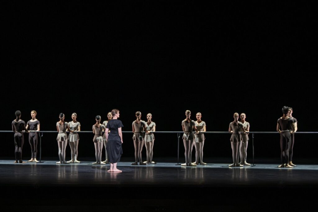 In review: 2026 summer performance at the royal opera house Dancers stand in a line at a barre that runs diagonally down the stage. Each dancer has another mirroring them, facing each other on either side of the barre. A light illuminates the centre, resulting in a reflection-like effect. Deborah stands barefoot in first position, watching the students at the barre. She wears a long black dress with short sleeves. The students wear beige leotards or dark brown unitards.
