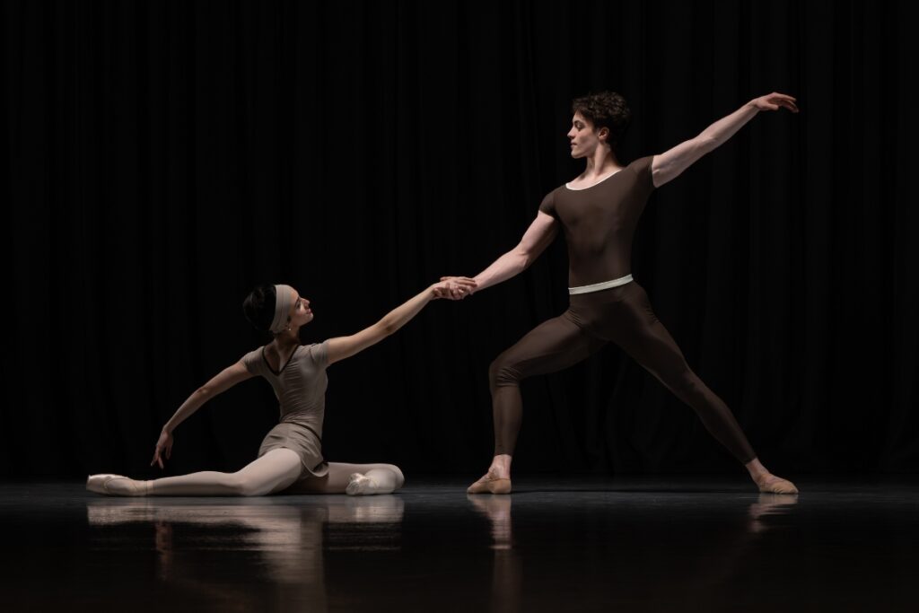 Alum louise flanagan on designing the costumes for cathy marston’s a line in reflection A girl sits on the ground facing stage left with her left leg bent in front of her and her right leg bent behind her. She raises her right arm up to hold the left hand of the boy across from her. He lunges towards her with his right leg front. His left arm is extended on a diagonal up behind him.