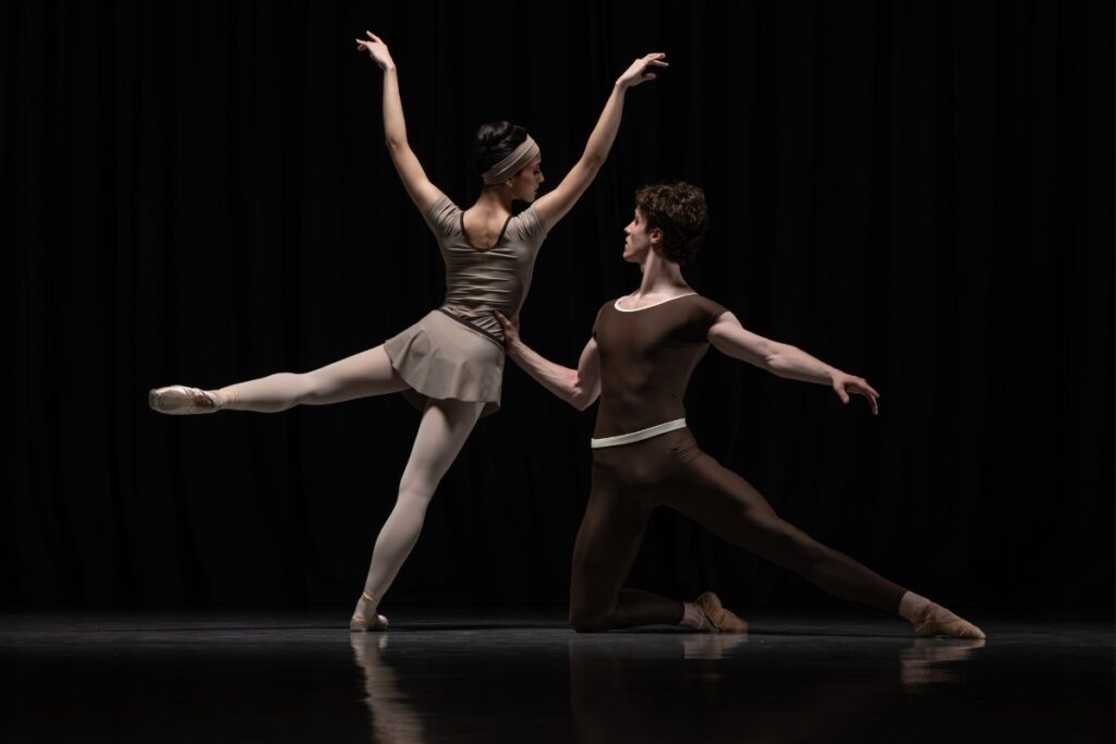 Alum louise flanagan on designing the costumes for cathy marston’s a line in reflection A girl in the beige leotard and skirt faces the back and leans off-balance on her right leg with her left leg in a parallel attitude to the side. Her arms are softly bent and raised above her head. A boy supports her waist with his right hand as he kneels on his right knee with his left leg extended to the back. His left arm is held to the back in a parallel line.