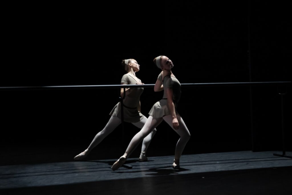 Two female dancers stand mirroring each other holding a barre. They lunge with the outside leg bent and forward with an extended leg to the back, both turned out. They tilt their heads away from the barre. They wear sepia grey leotards and wide headbands with pink tights and pointe shoes.