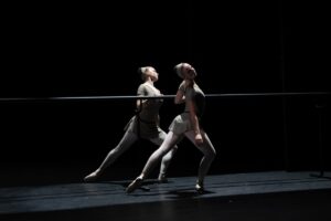Two female dancers stand mirroring each other holding a barre. They lunge with the outside leg bent and forward with an extended leg to the back, both turned out. They tilt their heads away from the barre. They wear sepia grey leotards and wide headbands with pink tights and pointe shoes.