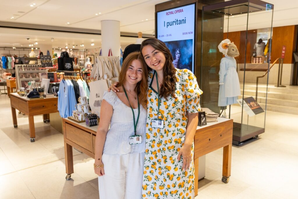 Behind the designs with centenary collection illustrators aimee tregunno and catja christensen Aimee and catja hold each other with one arm and smile at the camera. Catja leans her head down onto aimee's head. Aimee wears a white short sleeve top and white trousers. Catja wears a 3/4 sleeve white dress with oranges printed on it.