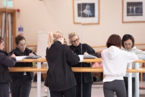 Three female teachers take notes while leaning over a barre. Their reflections are seen in the mirror in front of them.