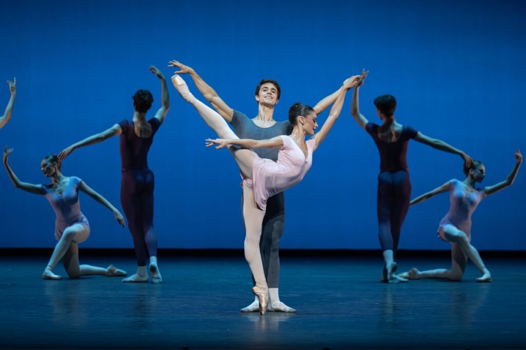 In review: 2026 summer performance at the royal opera house A young lady in a pink cap-sleeve leotard and matching short skirt poses in an arabesque on pointe with her right leg raised high behind her. A young man stands behind her in a charcoal grey unitard with both of his arms raised in a v above him. He holds her left hand in his own. He gazes up and smiles while she looks demurely downwards.