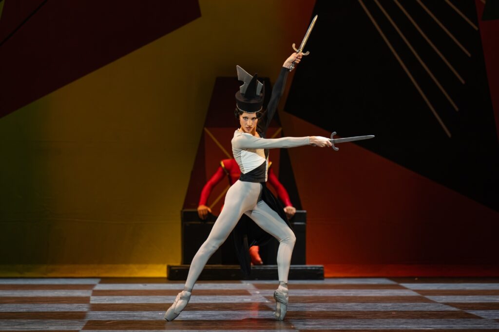 In review: 2026 summer performance at the royal opera house A young lady in a grey unitard with black and white colourblocked triangles on her top poses on pointe in a lunge to the left. Her left leg is bent to the front while her right leg is extended back. Her arms extend in a v to the side, each hand holding silver daggers. She stares intensely at the audience. She wears a chess piece inspired headpiece with angular black and grey sculptural shapes.