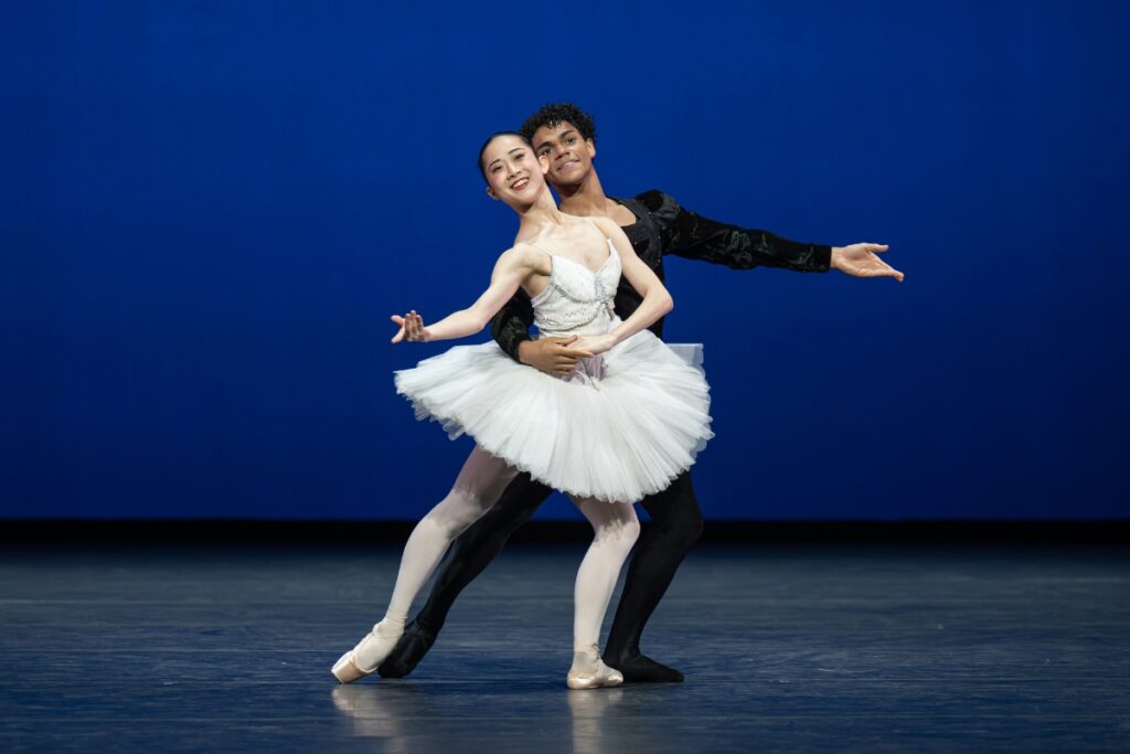 In review: 2026 summer performance at the royal opera house Two dancers lunge on a diagonal forward with their left legs bent to the front. The boy stands behind the girl, his right arm around her waist and her left hand holding his. Their other arms are softly extended to the side, curved upwards. They smile brightly to the audience. The girl wears a white pancake tutu with a bejeweled bodice. The boy wears black tights and a long sleeve top.