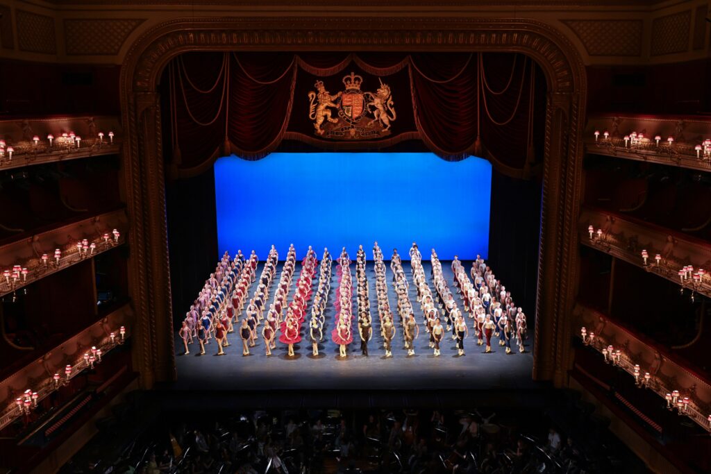 In review: 2026 summer performance at the royal opera house A wide angle view of the stage from above, with the tiers of seating framing the blue lit stage. 16 rows of students fill the stage, each with a different year group in uniform. They all stand in 5th position with their arms in low 5th and look up on a diagonal to the right.