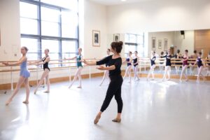 A female teacher dressed in a long-sleeve black shirt and black trousers with nude shoes demonstrates a tendu front to a group of young girls by the barre, who copy her pose.