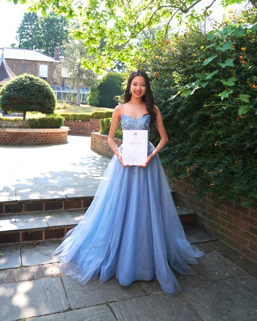 End-of-year celebrations and prize-giving ceremonies 2026 Juju holds her certificate in the shade of a green bush wearing a light blue flowy gown. Her long dark hair is straight and cascades over her shoulders.