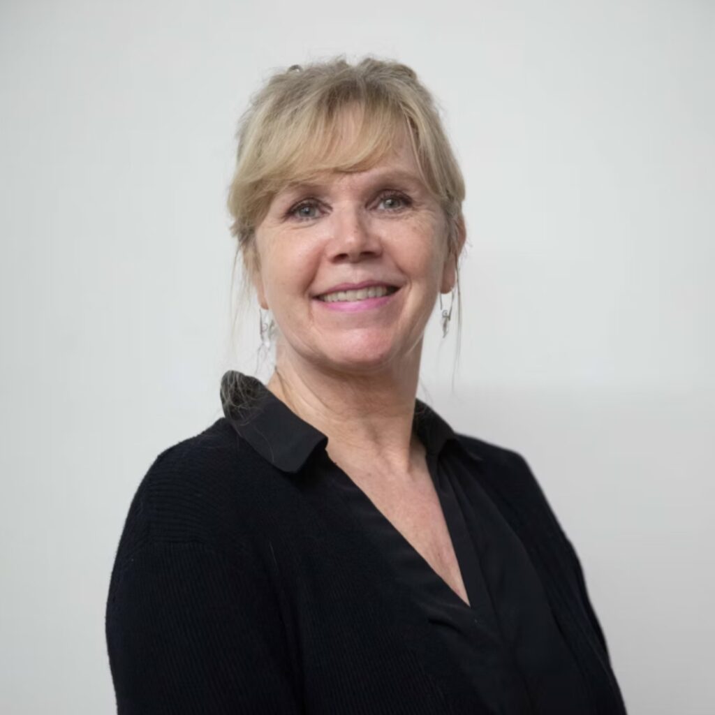 100 stories: karen paisey Karen wears a black v-neck shirt with her blond hair tied to the back. Her fringe falls gently to the right side of her face. She smiles at the camera.