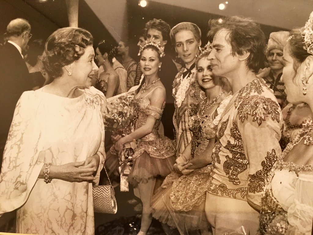 100 stories: karen paisey Karen paisey with queen elizabeth and rudolf nureyev in a group of dancers after performing. The photo has a sepia tone to it.