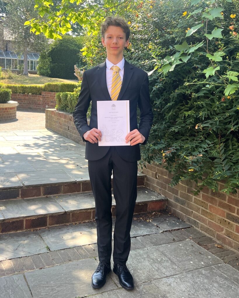End-of-year celebrations and prize-giving ceremonies 2026 Mathias wears a navy suit and yellow tie with black dress shoes. He holds his certificate in the shade of a green bush. His short brown hair is swept slightly to the side.