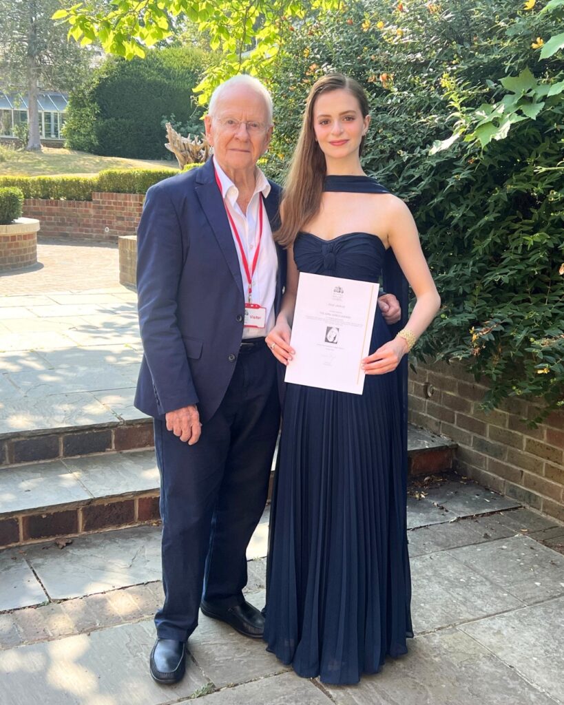 End-of-year celebrations and prize-giving ceremonies 2026 Nigel pegram wears a dark navy suit and stands next to jolie, who holds her certificate. Jolie wears a flowy navy strapless dress with a matching chiffon scarf. Her light brown hair is straight and falls on her shoulders.