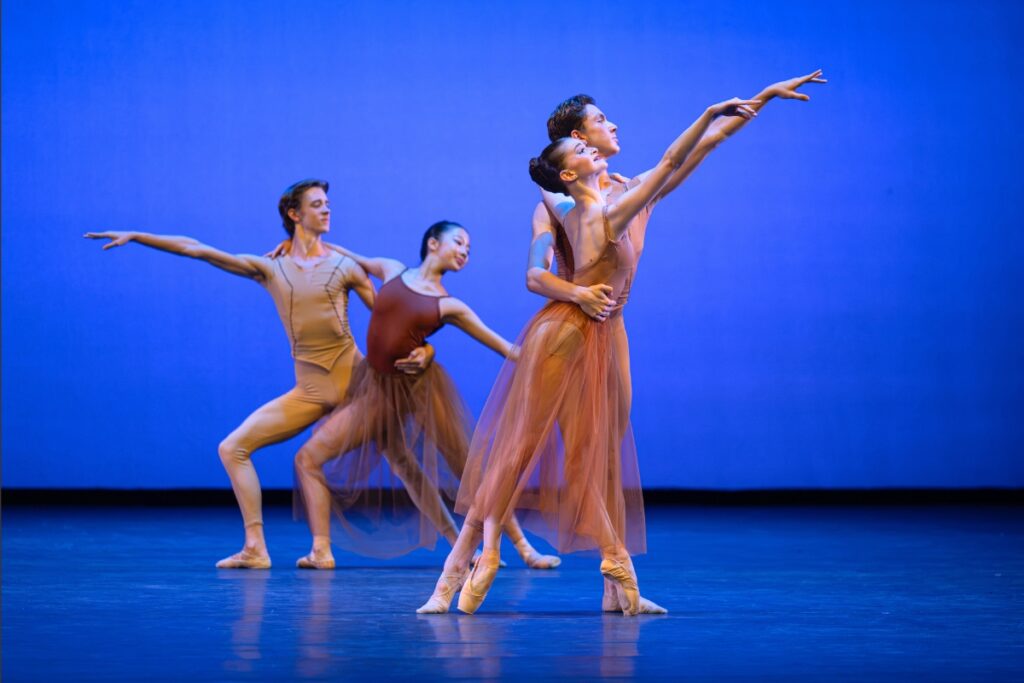 In review: 2026 summer performance at the royal opera house Four dancers wear shades of warm brown tones against a vibrant blue backdrop. One couple downstage poses in 4th position; the boy holds the girls waist from behind while they both reach up on a diagonal to the side. The girl is on pointe in skin-tone pointe shoes. She has a long tulle skirt and spaghetti strap leotard. The boy is in skin-tone shoes, brown tights and a brown short-sleeve shirt. The couple in the back lunges forwards on their right legs, with the boy holding the girl's waist from behind. Their arms are extended on a low angle to the side. The girl wears a dark brown leotard and tulle skirt. The boy wears a light mustard yellow short sleeve shirt and tights.