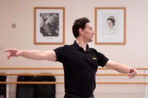 Valeri hristov wears a black polo shirt as he demonstrates a step to the class. The photo is cropped from his waist up. In the background are portraits of princess margaret and sir frederick ashton.