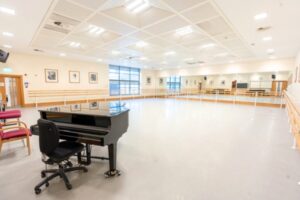 A wide angle view of the bright, spacious princess margaret studio in upper school. A piano is in the foreground, and a large window is adjacent to the wall of mirrors. Portraits line the walls in wooden frames, matching the warm wood baseboards.