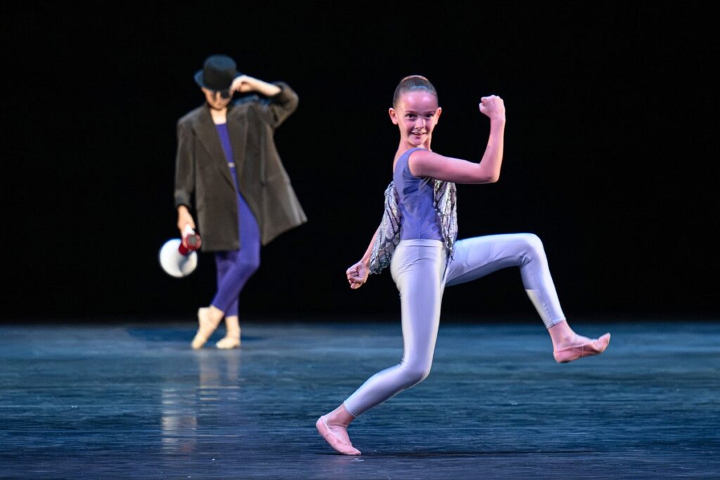 In review: 2026 summer performance at the royal opera house In the foreground, a young girl in light grey leggings, a purple leotard and iridescent insect wings on her front and back stands on a bent right leg with her left leg bent at a 90 degree angle. Her right arm is raised front in a similar 90 degree angle. Her left arm hangs loosely to the side. In the background upstage, a young boy stands on his right leg with his left foot crossed over. He wears an oversized coat, indigo tights and a black bowler hat. He tips his hat forward with his left hand and holds a white and red megaphone in his right hand, hanging down by his side.