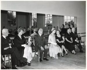 The guests and faculty sit in the front of the studio in front of the mirror facing a classroom of young girls in white leotards.