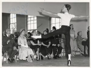 The guests and faculty sit in the front of the studio in front of the mirror facing a classroom of young boys in white shirts and black tights with white socks and black shoes. The camera is in the back of the studio, capturing a student mid jump with his left leg in a la second and his arms held to the side.