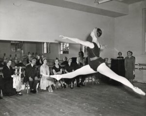 The guests and faculty sit in the front of the studio in front of the mirror facing a classroom of young girls in black leotards. The camera is in the back of the studio, capturing a young dancer mid grand jete with her right leg and left arm forward.