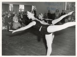 The guests and faculty sit in the front of the studio in front of the mirror facing a classroom of young girls in black leotards. The students penche forward with their left legs behind in arabesque. The camera angle is from the back of the studio.