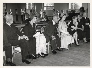 The guests and faculty sit in the front of the studio in front of the mirror facing a classroom of young girls in white leotards, who are seen in the mirror reflection.