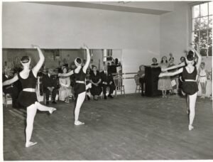 The guests and faculty sit in the front of the studio in front of the mirror facing a classroom of young girls in black leotards. The camera is in the back of the studio, showing the students in a develope front with their left legs. Their right arms are raised in fifth and their left arms are held in second.
