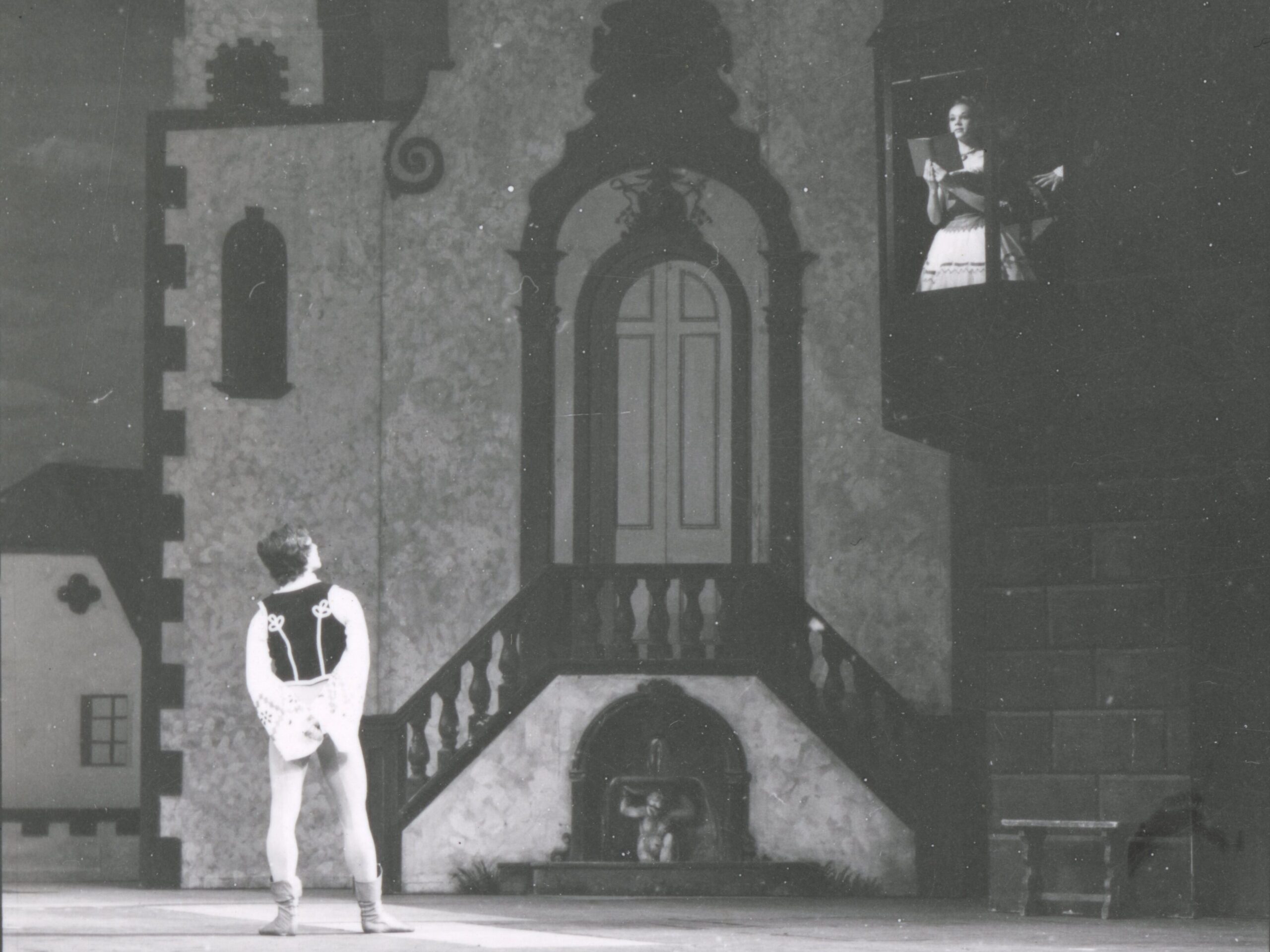 100 stories: karen paisey Karen reads a book as coppélia in a school production. She sits high on a balcony. Below her, a male dancer gazes up at her.