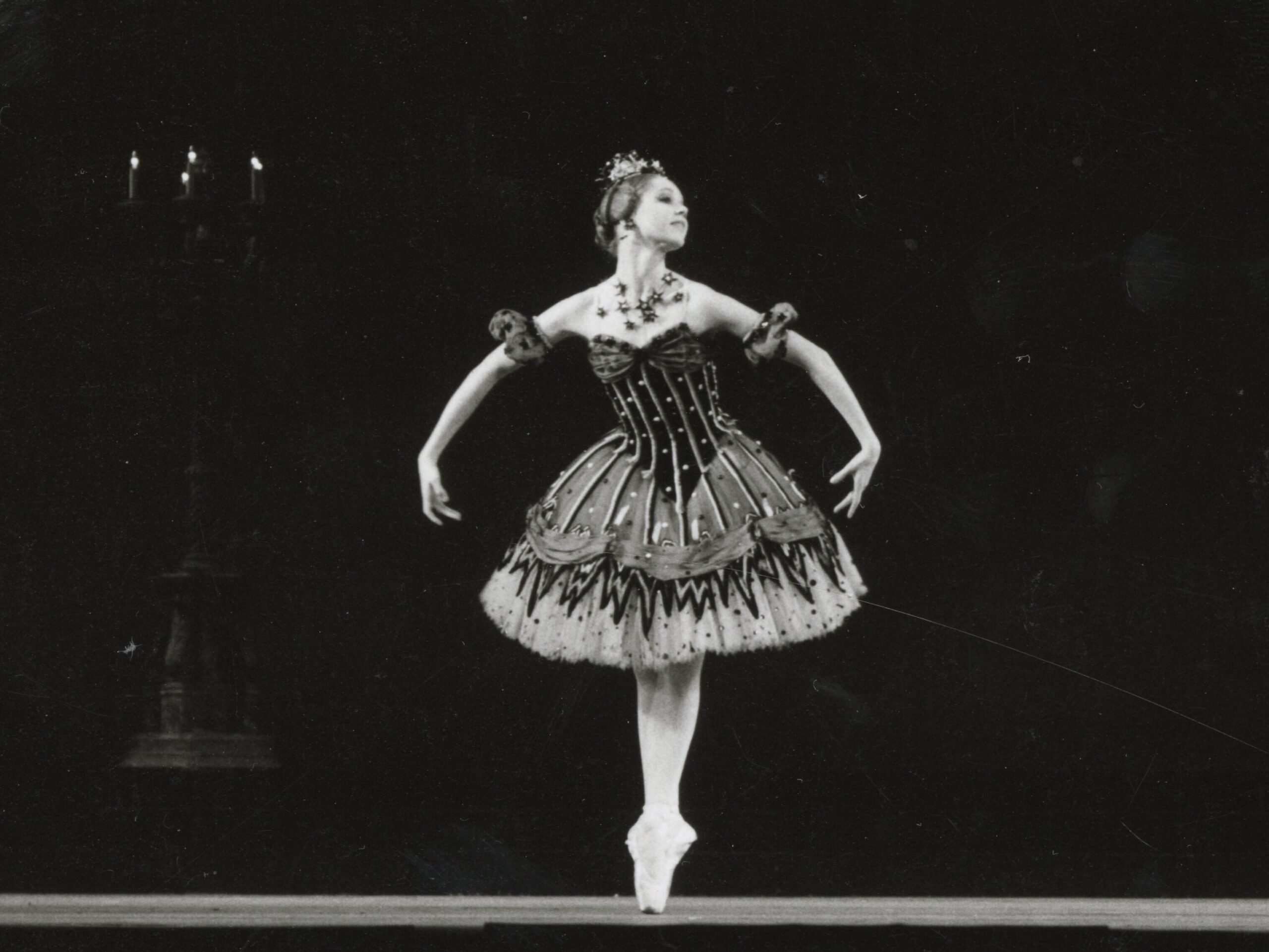 100 stories: karen paisey Karen wears a knee-length black tutu as she balances in 5th position on pointe. Her arms are curved low to her sides. Her head is turned far to the left, looking over her shoulder to the audience.