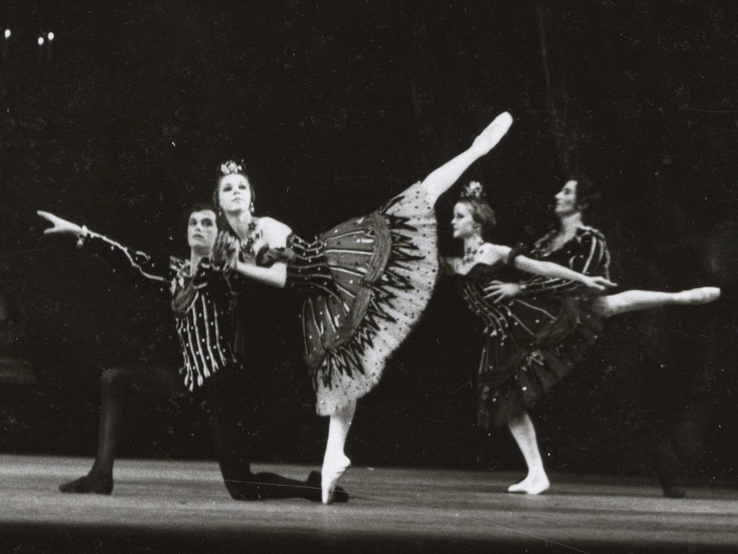 100 stories: karen paisey Karen penches on pointe while holding the arm of a kneeling male dancer in front of her. She wears a knee-length black tutu.