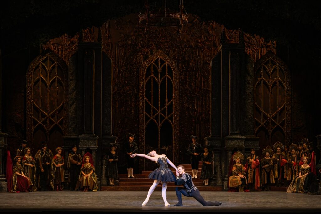 In review: 2026 summer performance at the royal opera house A wide shot of the stage. Centre: the black swan arches back with her right arm straight and next to her head, which gazes straight up. Her right leg is in tendu back. She holds siegfried's hand. With her left as he kneels in front of her. He puts her hand to his cheek.