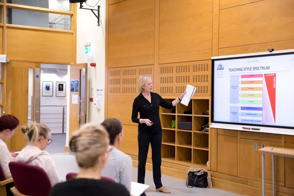 Ballet teaching qualifications in the uk explained A teacher dressed in all black gestures towards a projector screen with the slide title reading 'teaching style spectrum. ' in the foreground, other teachers sit in chairs facing the screen while taking notes.