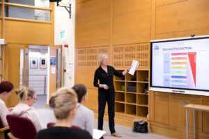 A teacher dressed in all black gestures towards a projector screen with the slide title reading 'teaching style spectrum. ' in the foreground, other teachers sit in chairs facing the screen while taking notes.