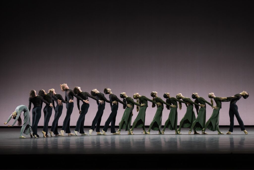 In review: 2026 summer performance at the royal opera house Students stand holding each others waist in a curving line. Most face stage left and tilt their heads back. They stand in height order from shortest to tallest, which creates a slight curve up. At the far left, a girl bends her back deeply with her arms above her head. On the right, a boy faces the group and arches back. They all wear loose trousers and long sleeve shirts, the boys in black and the girls in olive green.
