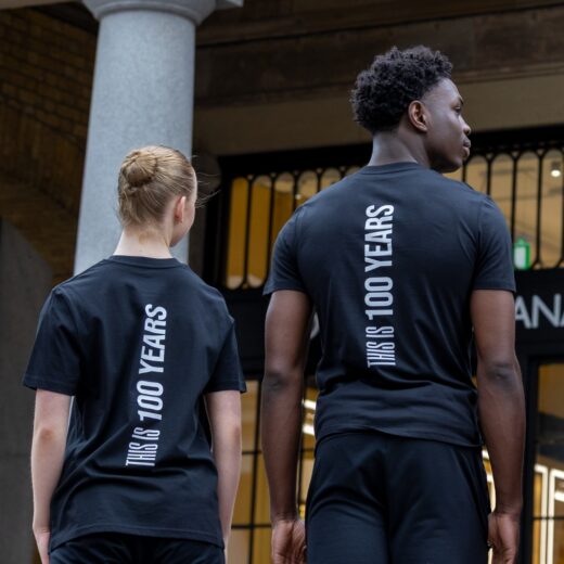 Product image of 'This is 100 years t-shirt' in black. Worn by Royal Ballet School students in Upper School, Covent Garden. A collection of merchandise in celebration of 100 years of The Royal Ballet School.