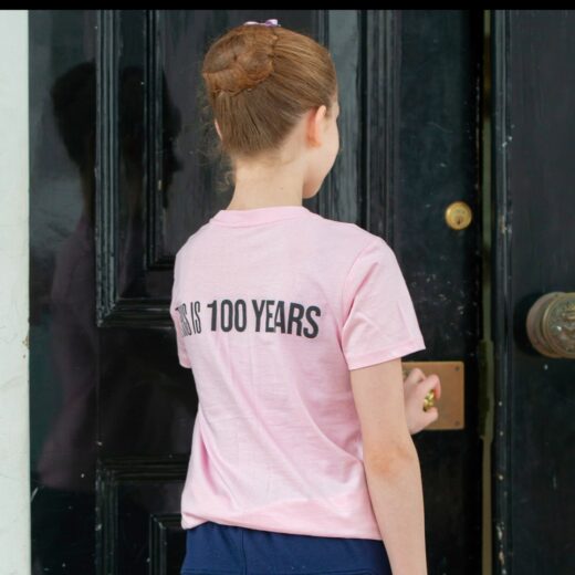 Product image of 'This is 100 years children's t-shirt' in pink. Worn by a Royal Ballet School student at White Lodge. A collection of merchandise in celebration of 100 years of The Royal Ballet School.