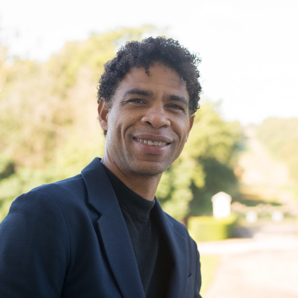 Carlos acosta smiles at the camera in front of the back garden at white lodge. He wears a black crew neck shirt and a black jacket.