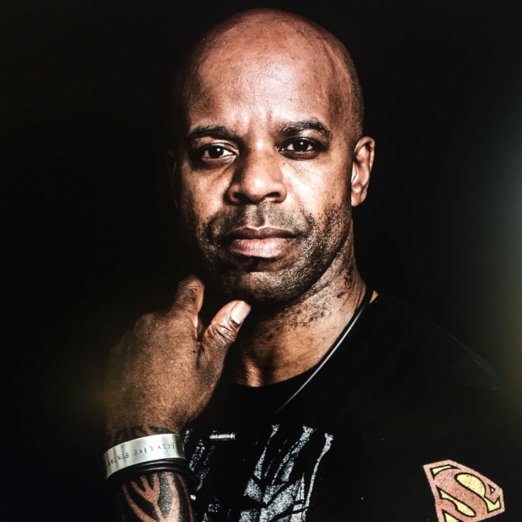 Denzil slightly smiles at the camera with his left hand under his chin. He wears a black shirt with a superman logo on the left shoulder and a silver watch.