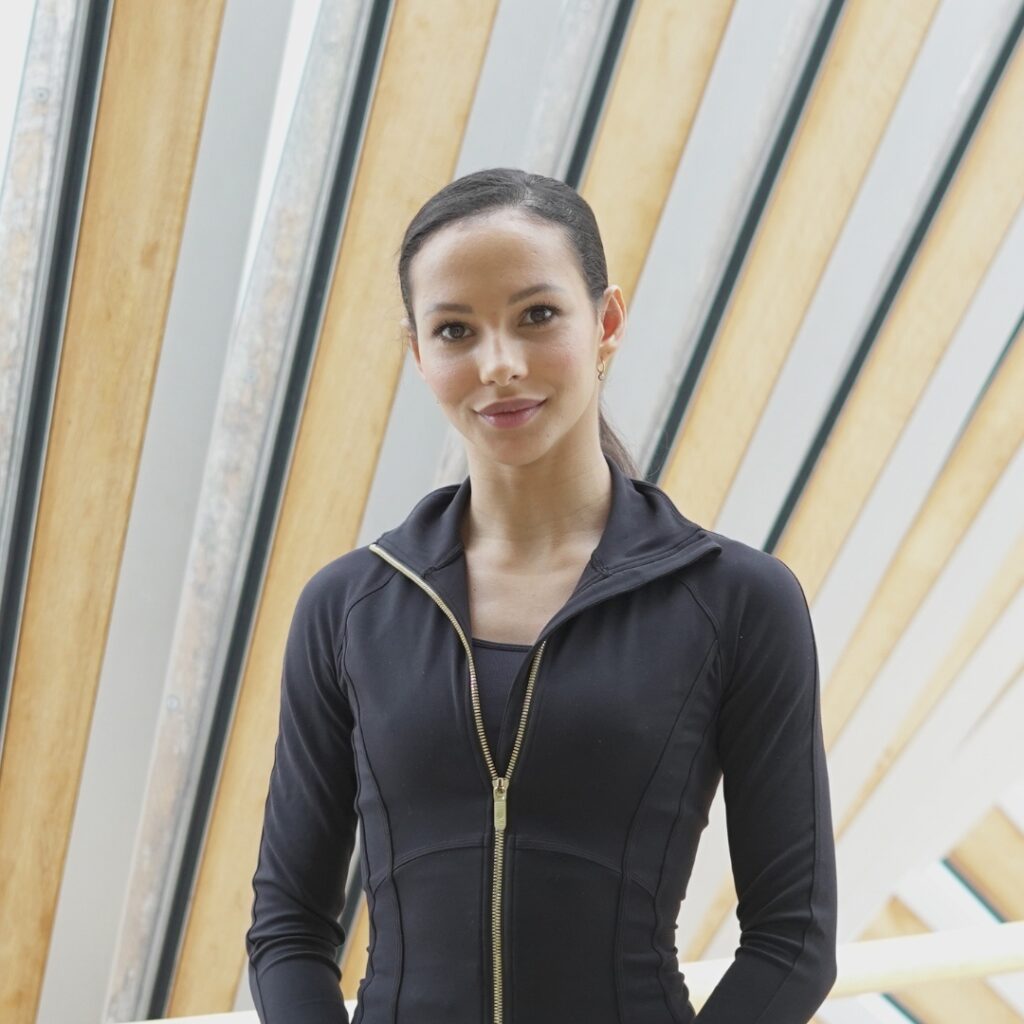 Francesca hayward stands on the bridge of aspiration with her hair tied back in a ponytail. She wears a long sleeve black jumper with a gold zipper partially undone with a black undershirt.