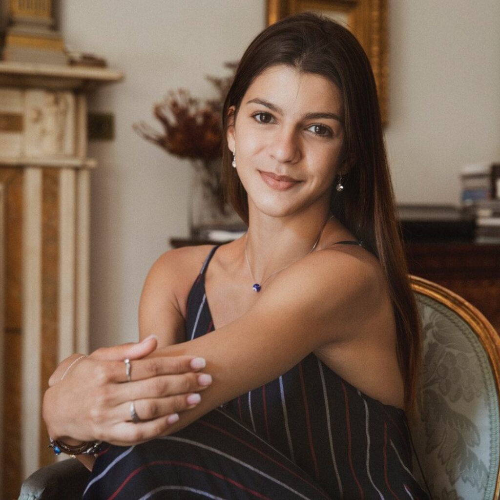 Mayara sits in a chair with her long straight hair falling down her back. She wears a spaghetti strap black jumpsuit with thin white stripes. She holds her left elbow with her right hand and props both on her left knee, which is crossed on top of her right leg.