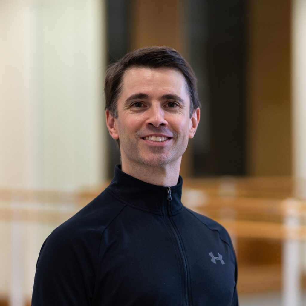 Ricardo smiles at the camera in a ballet studio with wooden barres blurred in the background. He wears a high neck, long sleeved black shirt.