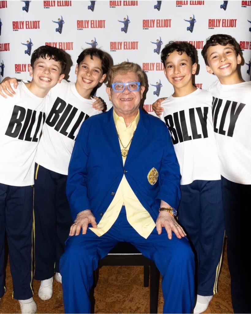 Year 8 student rafferty on starring as the titular role in billy elliot the musical The four billys surround elton john, who sits on a chair in a cobalt blue suit and butter yellow shirt. They stand in front of a wall that has the billy elliot the musical logo printed repeatedly.