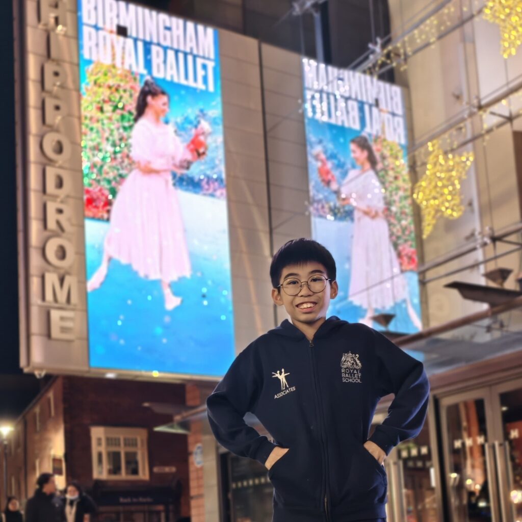 Mid associate students share what they love about the associate programme Charles smiles in front of a birmingham royal ballet 'the nutcracker' poster. He wears an associates tracksuit with his hands on his hips.