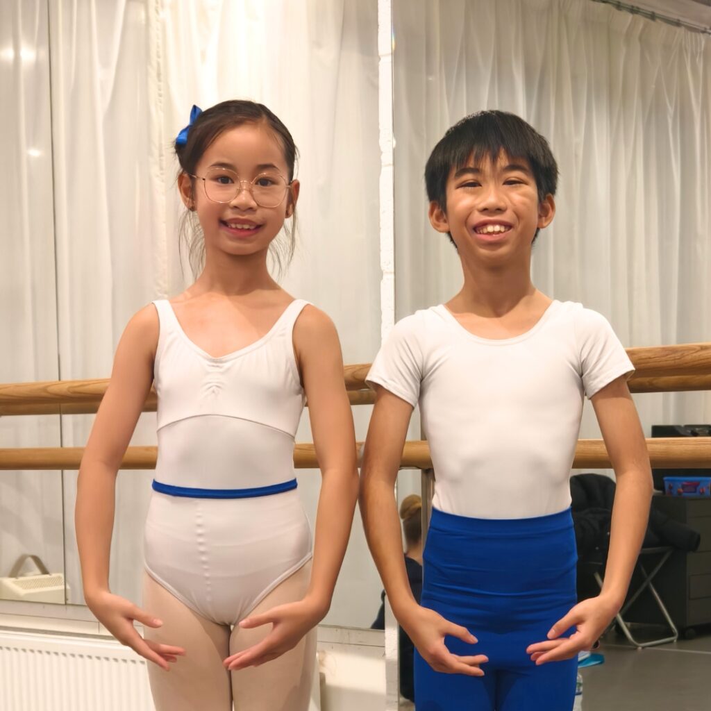 Mid associate students share what they love about the associate programme Charles stands to the left of his sister, charlotte, in junior associate uniforms in a studio. Charles wears a white t-shirt and blue tights. Charlotte wears a white leotard with a blue belt.