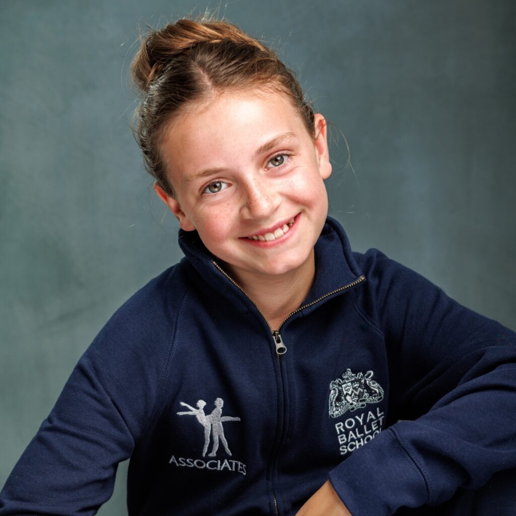 Mid associate students share what they love about the associate programme Grace smiles at the camera in front of a blue-grey backdrop. Her hair is in a bun and she wears an associates tracksuit.