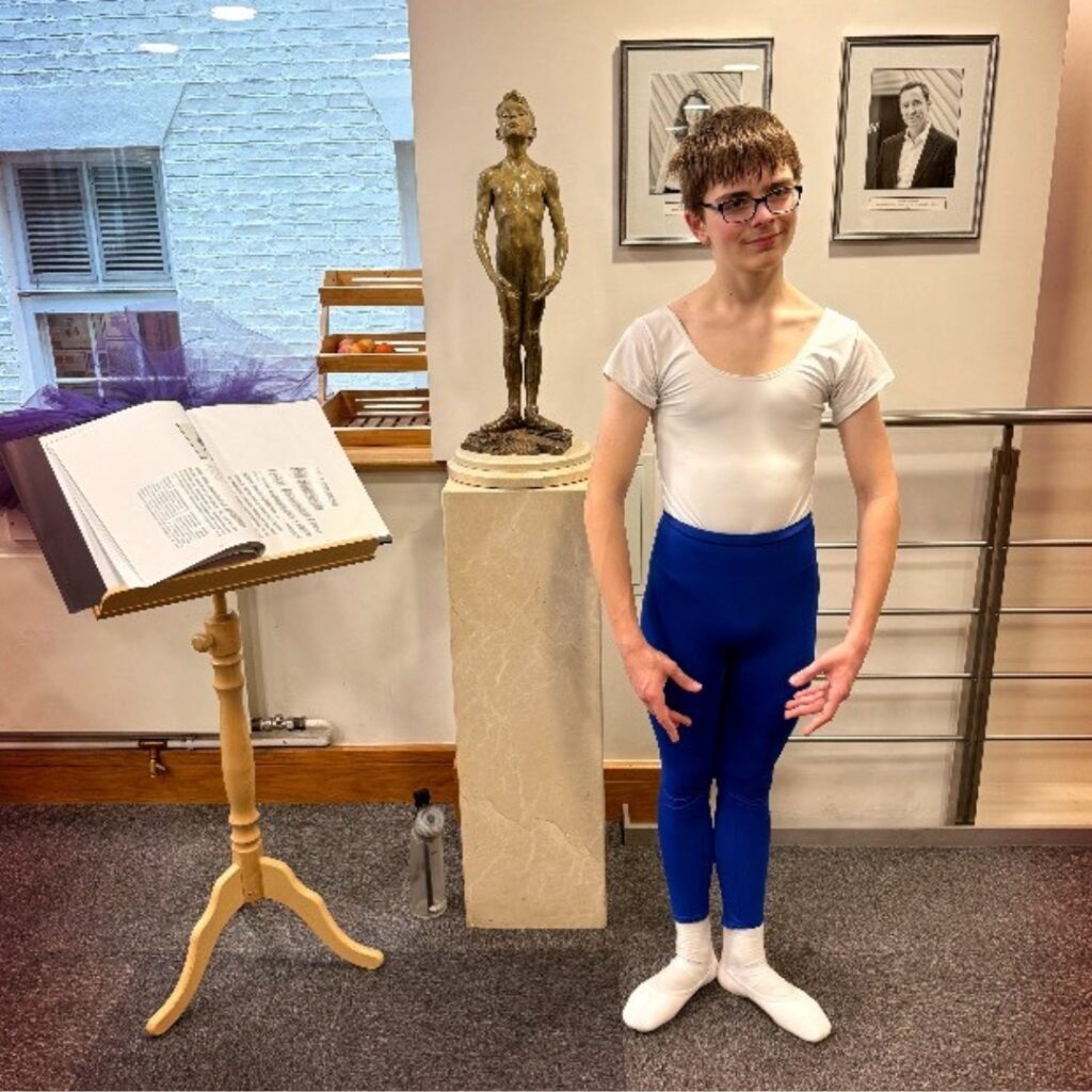 Mid associate students share what they love about the associate programme Joshua stands in first position in the foyer at upper school. He wears a white shirt, blue tights, white socks and shoes, and glasses.