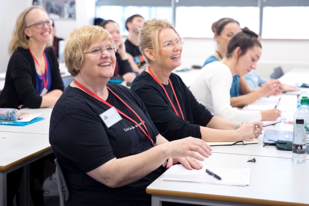 Affiliate teacher spotlight: julia barry Julia barry sits in the foreground at a desk, smiling at the speaker. Behind her are rows of other smiling dance educators.